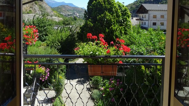 A balcony with a view of a mountainous landscape, a garden with red flowers, and a clear blue sky.