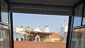 Een open balkon met een tafel en twee stoelen, uitzicht op een gebouw met rode pannendaken, en een heldere lucht.