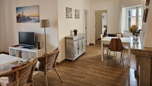 Een ruime woonkamer met een televisie, rieten stoelen, een eettafel en een keuken met een raam.