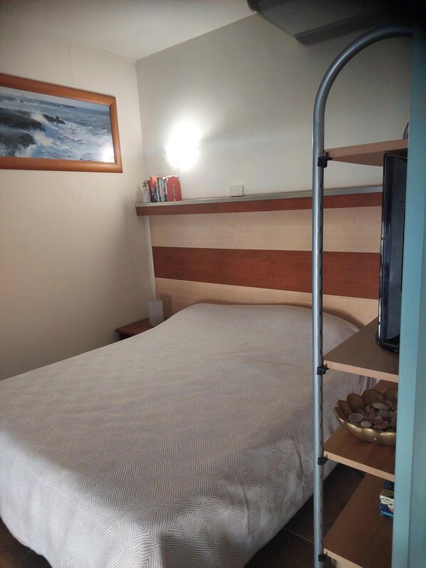 Ett hotellrum med en våningssäng, en säng med ett mönstrat överkast, en hylla med böcker och en liten växt, och en tavla på väggen.