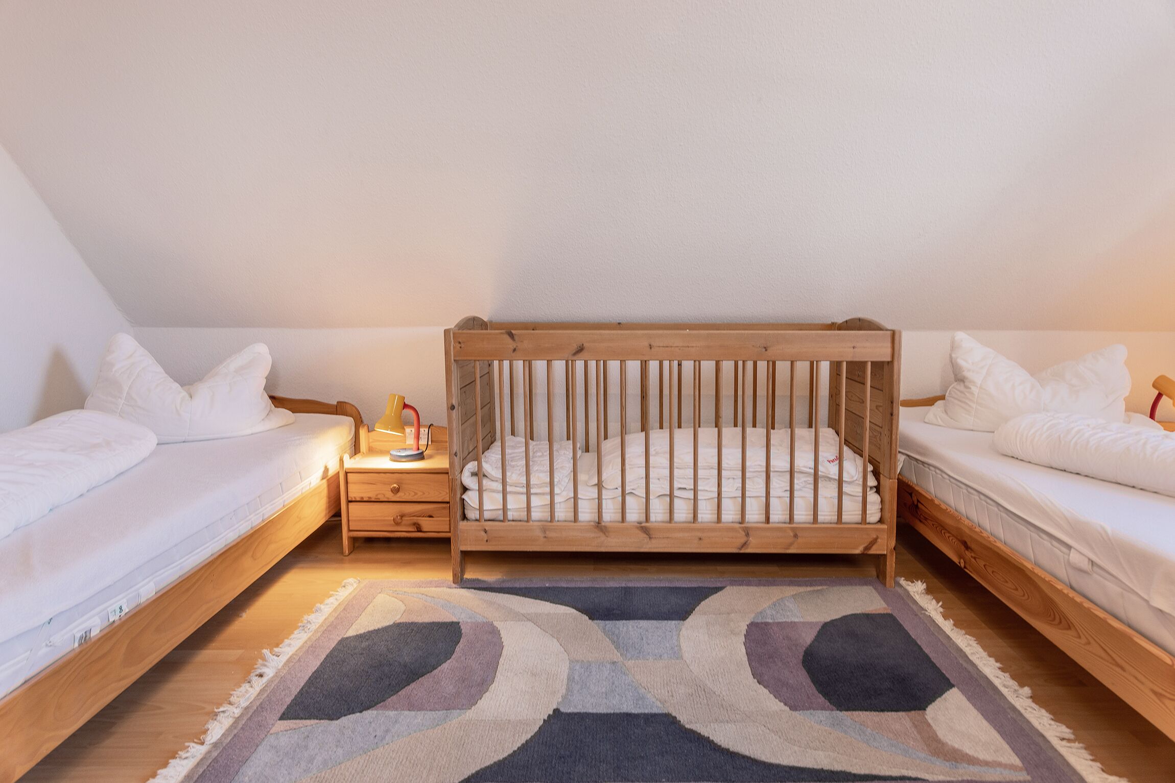 A bedroom with a wooden crib, two single beds, a nightstand, and a patterned rug.