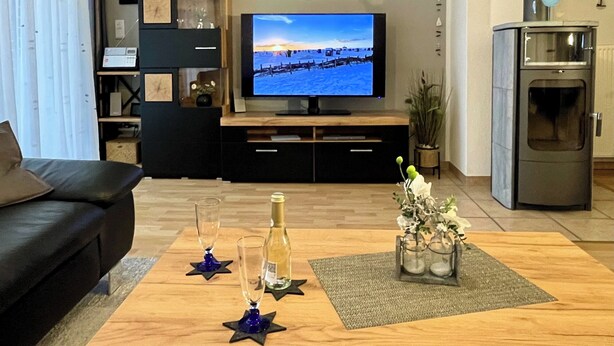 A living room with a flat-screen TV, a black sofa, a wooden coffee table with decorative items, and a kitchen area with a built-in oven. - Generous and tasteful: This terraced house of about (Carolinensiel)