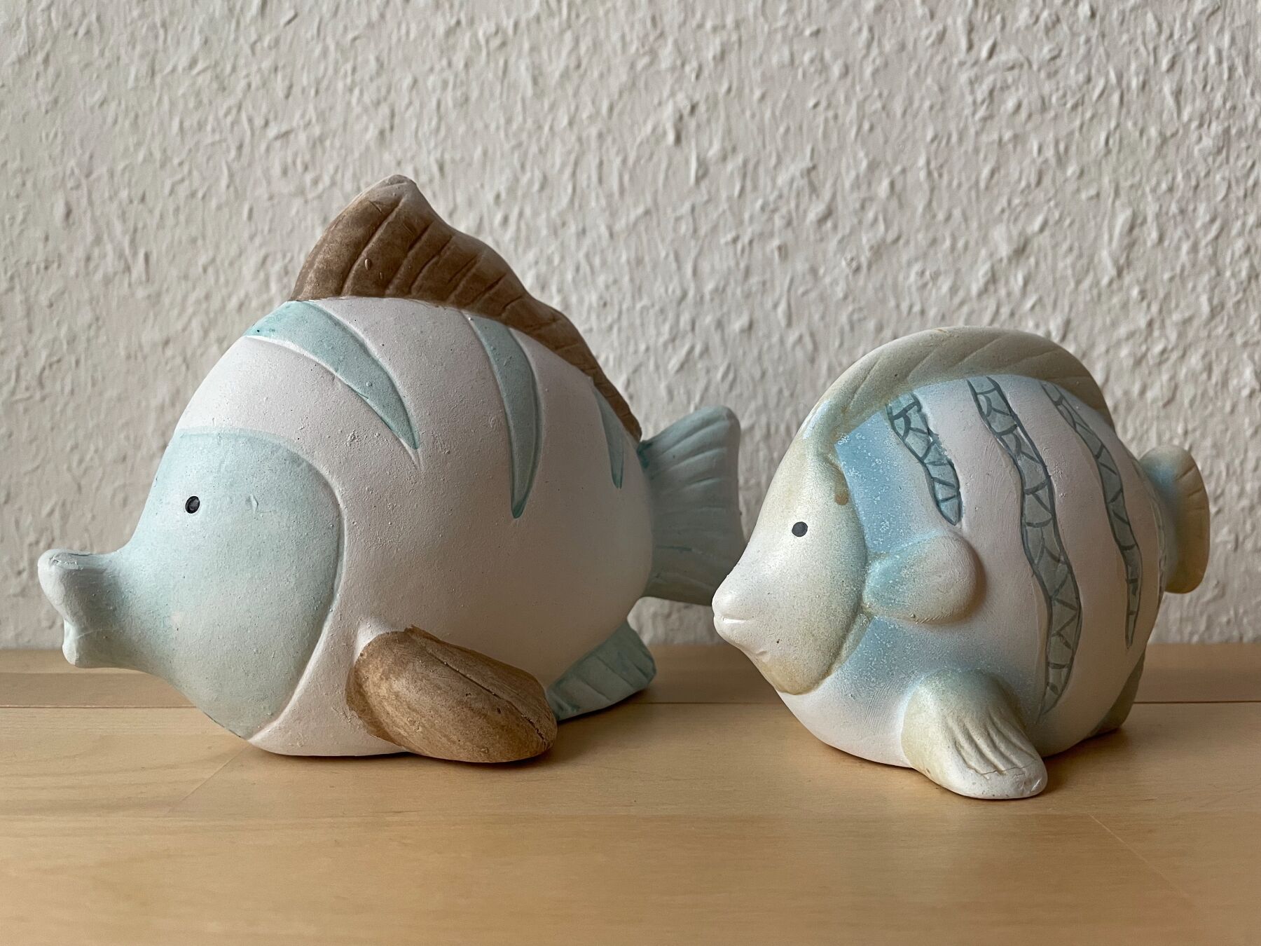 Two ceramic fish figurines on a wooden surface against a textured wall.