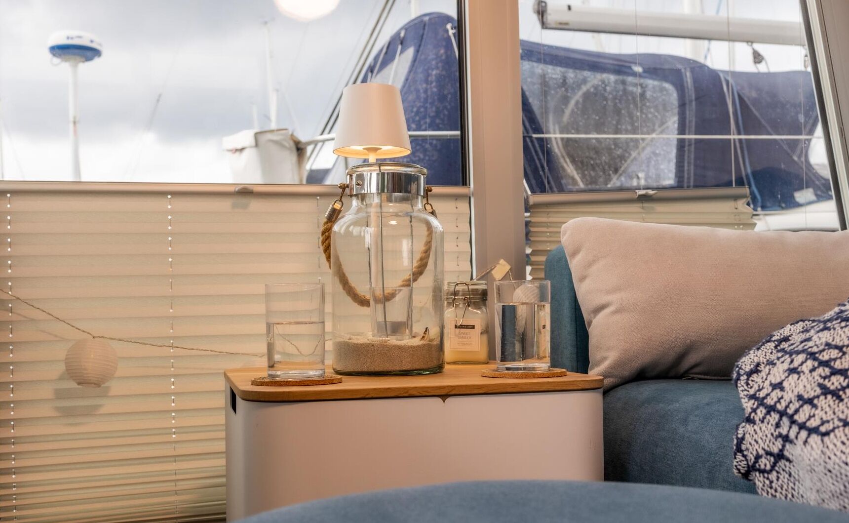 A cozy interior space with a wooden side table holding a glass jar with a rope handle, a candle, and a glass of water. A cushioned bench and a window with blinds are also visible.