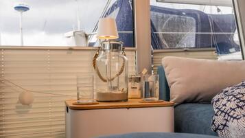 A cozy interior space with a wooden side table holding a glass jar with a rope handle, a candle, and a glass of water. A cushioned bench and a window with blinds are also visible.