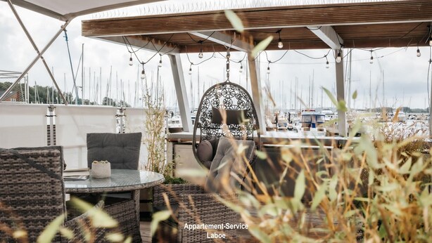 A covered outdoor seating area with wicker furniture, a hanging chair, and string lights. - Houseboat Canoga in Labo marina (Laboe)