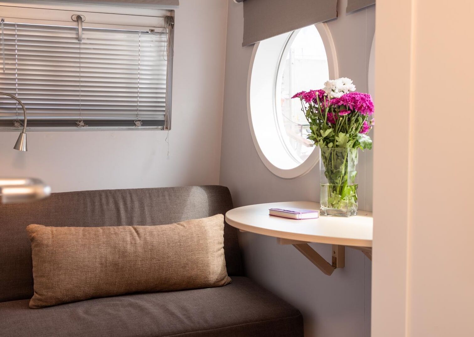 Ett rum med en brun soffa, ett litet runt bord med en vas med blommor, ett fönster med persienner och en lampa.