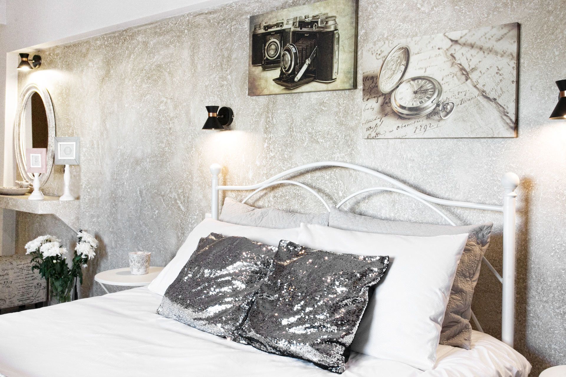 A neatly made bed with white linens and a black sequined pillow, a white headboard, and a wall with a vintage camera and a clock.