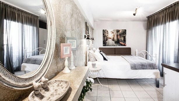 A bedroom with a large mirror, a bed with white bedding, a small table with a lamp, and a painting on the wall. - Asprolithos Studios (Skiathos)
