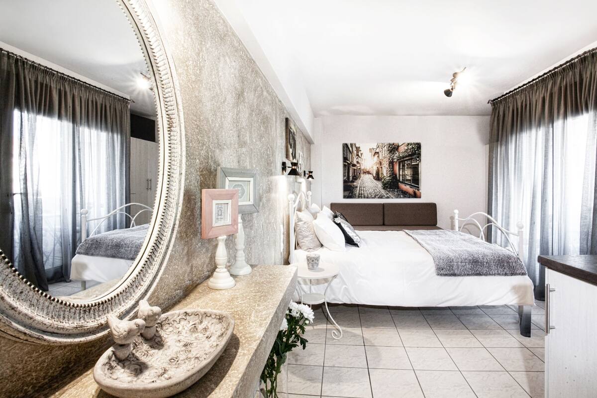 A bedroom with a large mirror, a bed with white bedding, a small table with a lamp, and a painting on the wall.