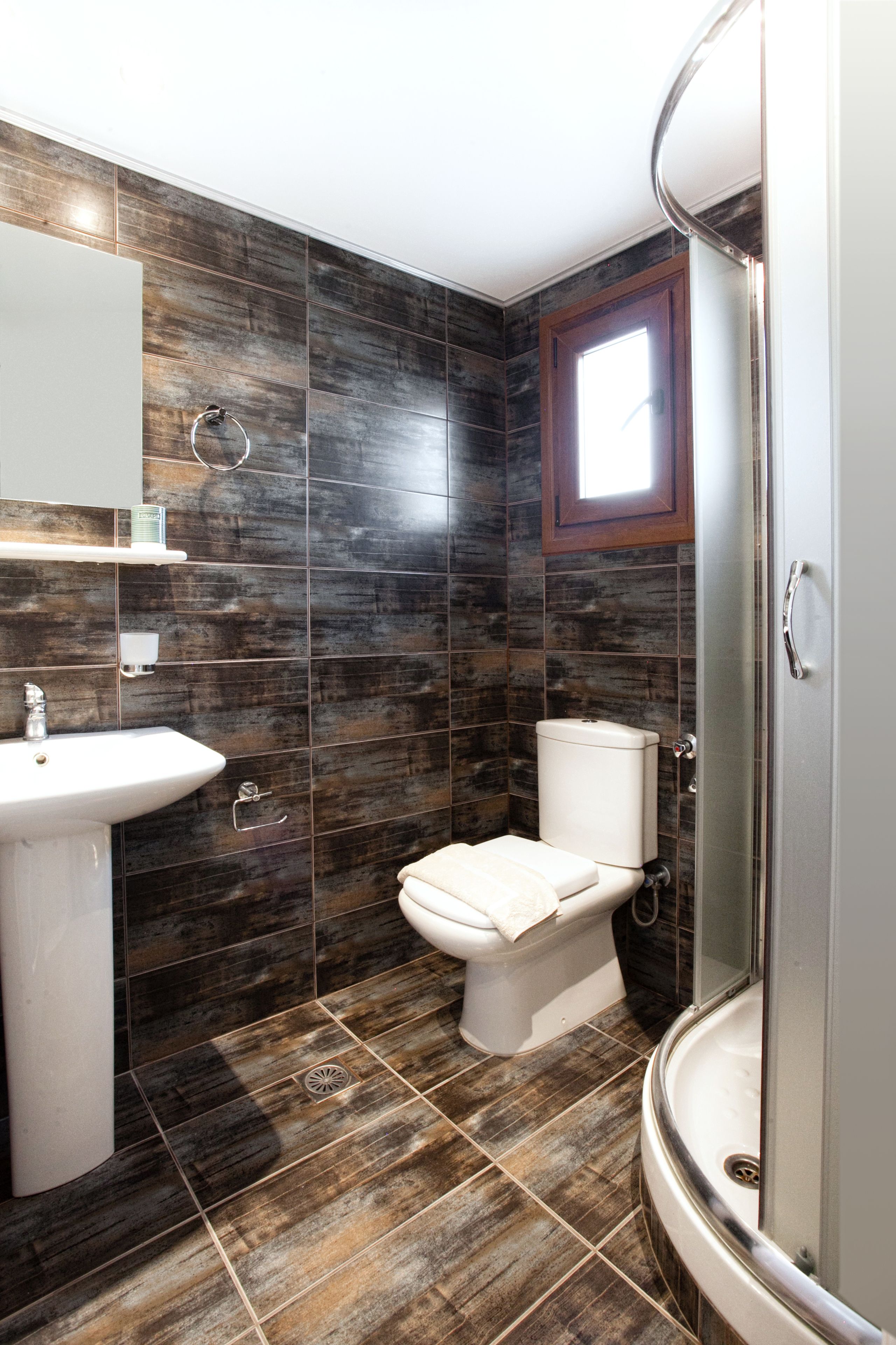 A modern bathroom with dark wood-patterned tiles, a white sink, a toilet, and a glass-enclosed shower.