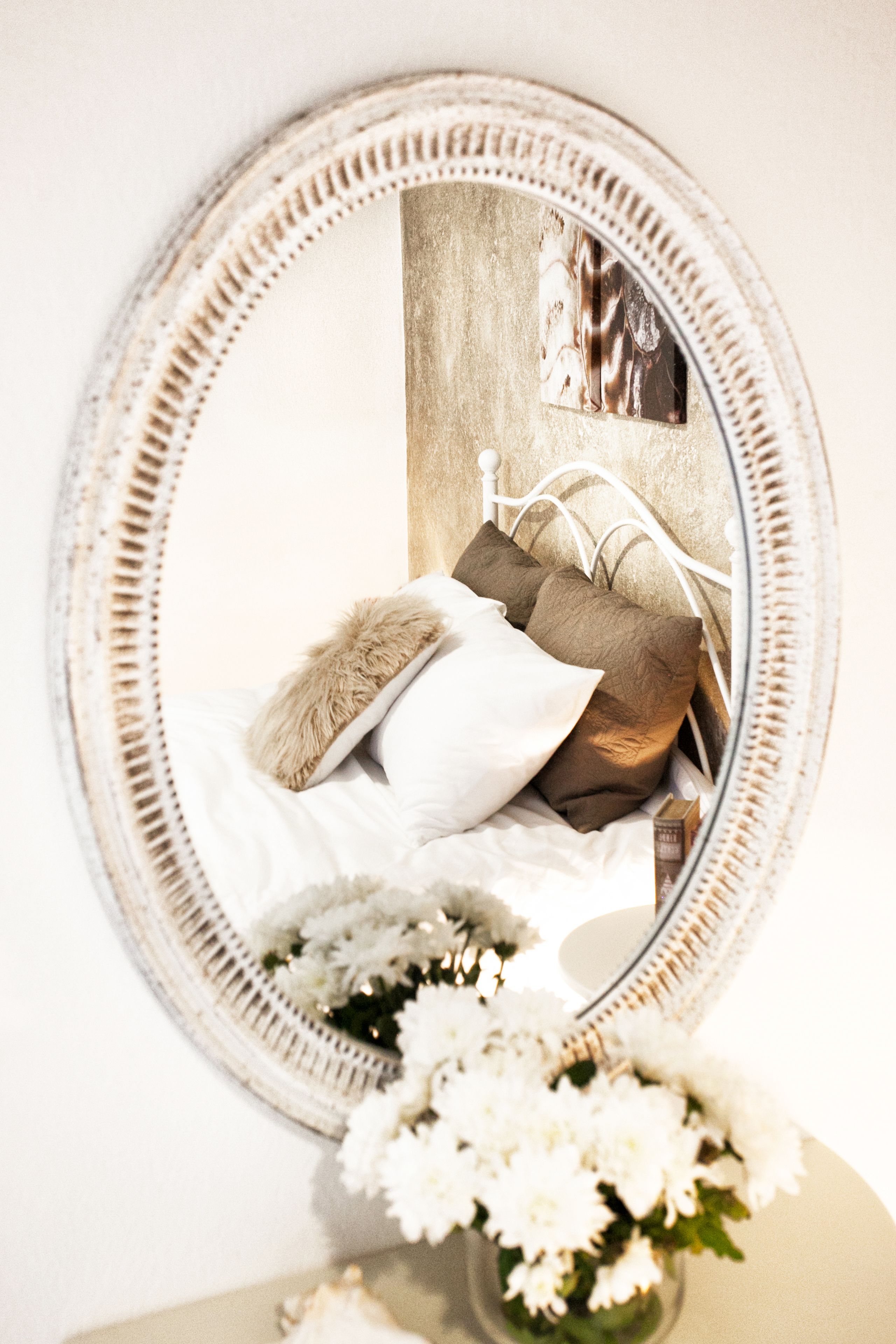 A round mirror with a decorative frame reflecting a bed with white and brown bedding, a fluffy pillow, and a vase with white flowers.