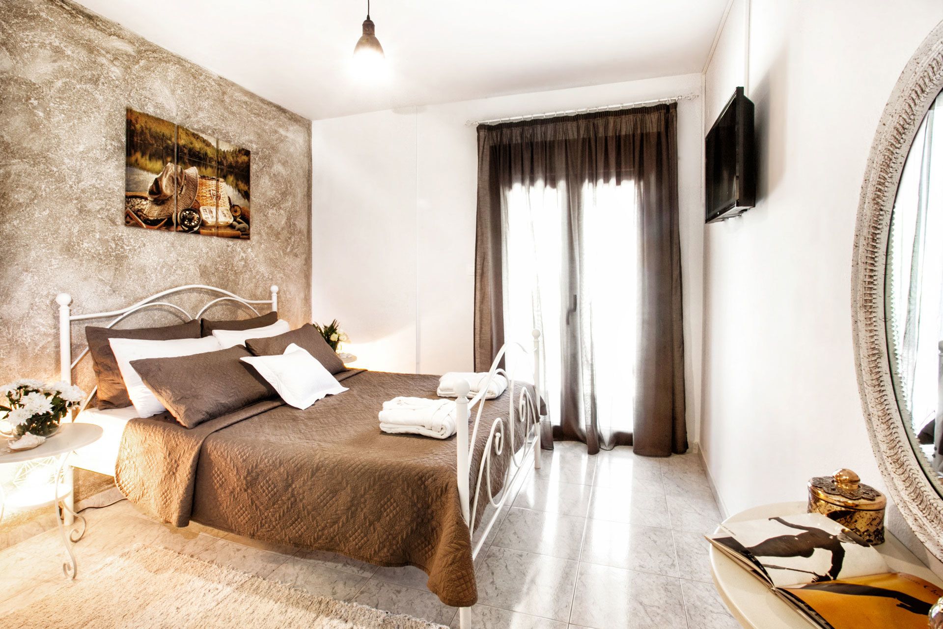 A bedroom with a bed, a bedside table, a mirror, a painting on the wall, and a window with curtains.