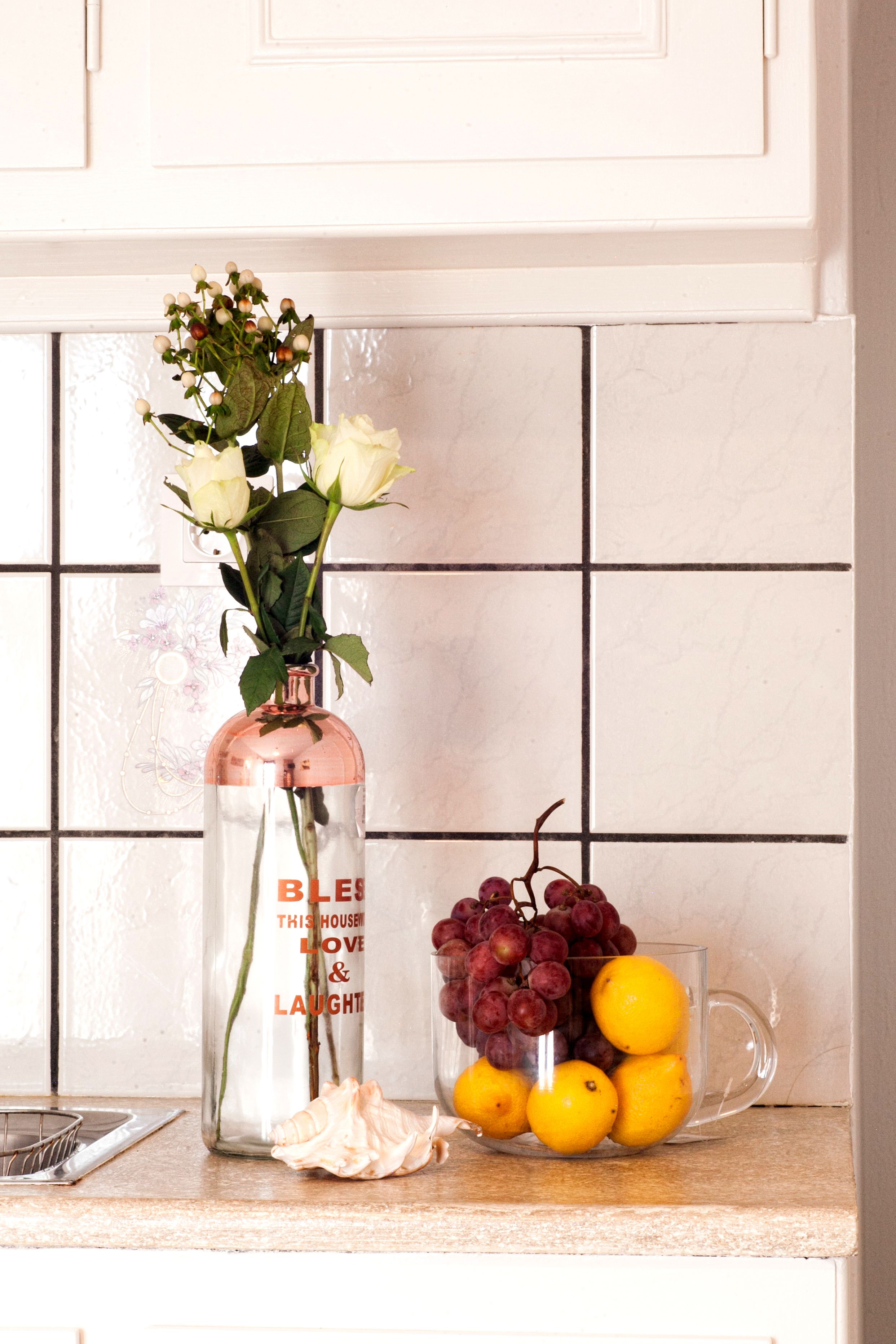 Una encimera de cocina con un florero transparente que contiene rosas blancas y una taza de vidrio con limones y uvas.