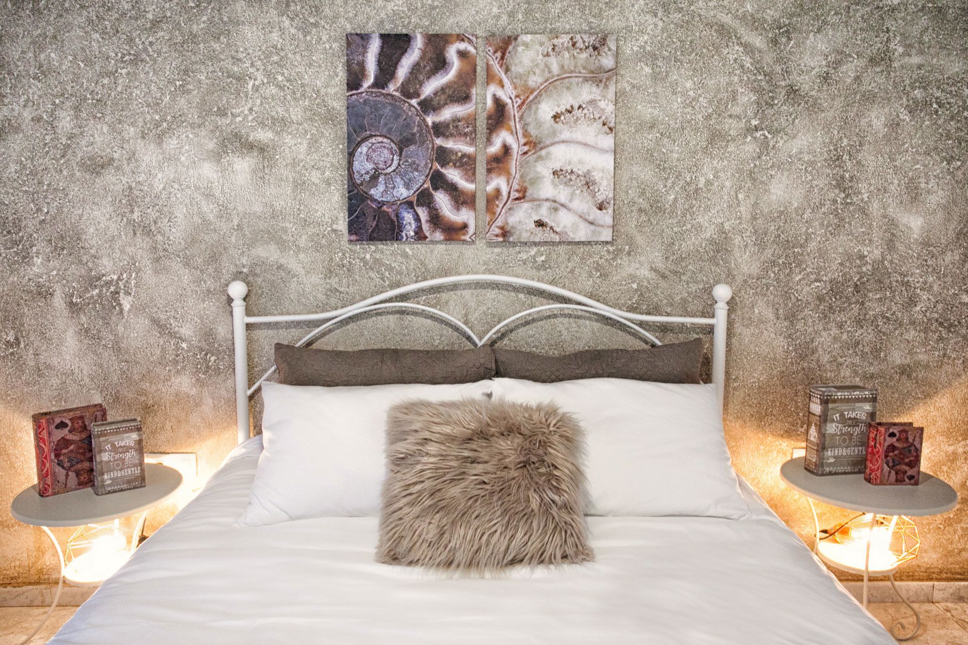 Una cama con cabecera blanca, un cojín mullido y un colchón blanco. Hay dos mesitas de noche con libros y una lámpara. La pared detrás de la cama tiene un diseño texturizado.