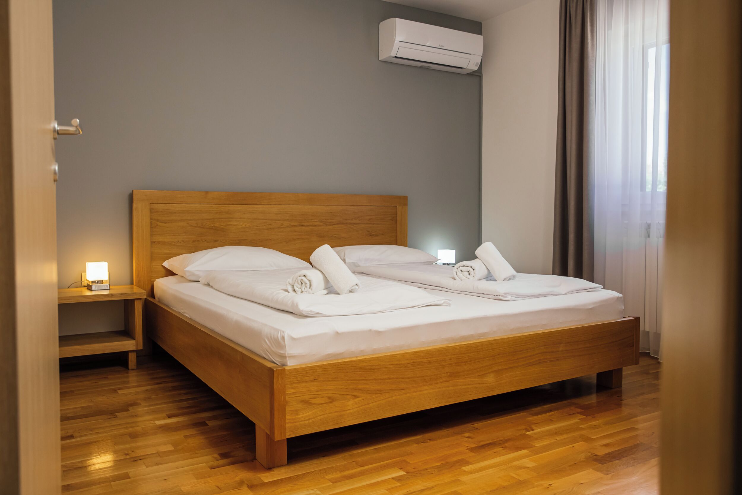 A neatly made bed with white linens and towels, a wooden headboard, a bedside table with a lamp, and an air conditioning unit on the wall.