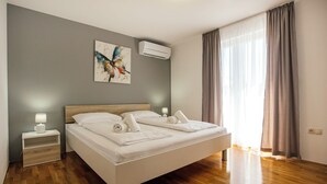 Moderni hotellihuone, jossa on puulattia, valkoisilla lakanoiden peitetty sänky, yöpöytä lampun kanssa, taulu seinällä ja ikkuna, jossa on verhot.