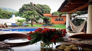 Relax in a serene garden setting with a pool, hammock, and dining area amidst lush greenery and a traditional building.