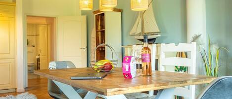 A dining area with a wooden table, white chairs, a wicker basket with fruits, a bottle of wine, and a sailboat decoration.