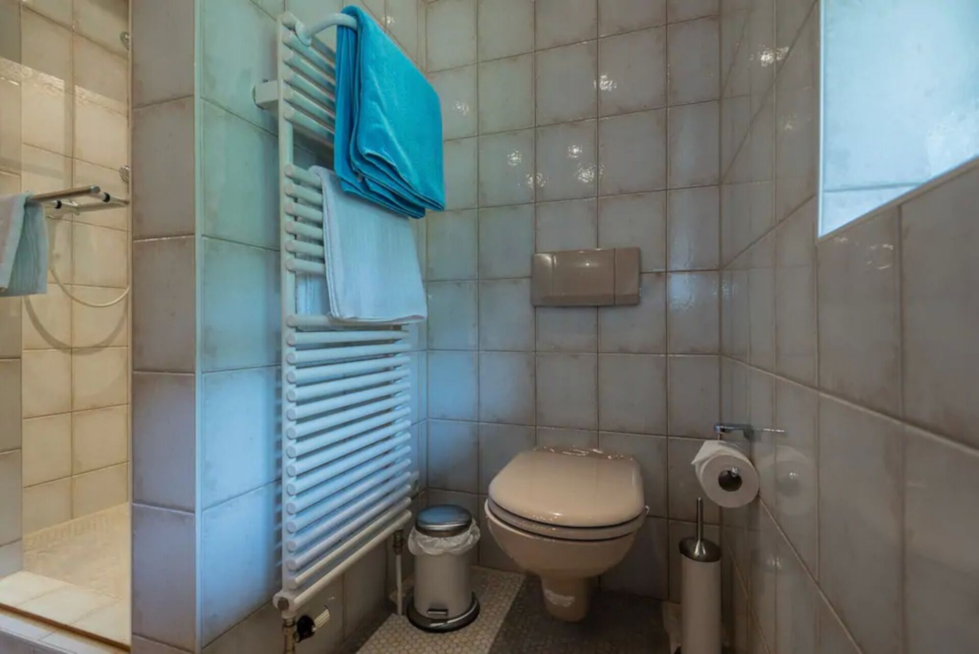 A small bathroom with a toilet, a radiator, a trash bin, and a towel rack with a blue towel.