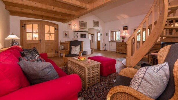 A spacious living room with a high wooden ceiling, a red sofa, a wicker chair, and a staircase. - Farmhouse Tegernsee (Warngau)