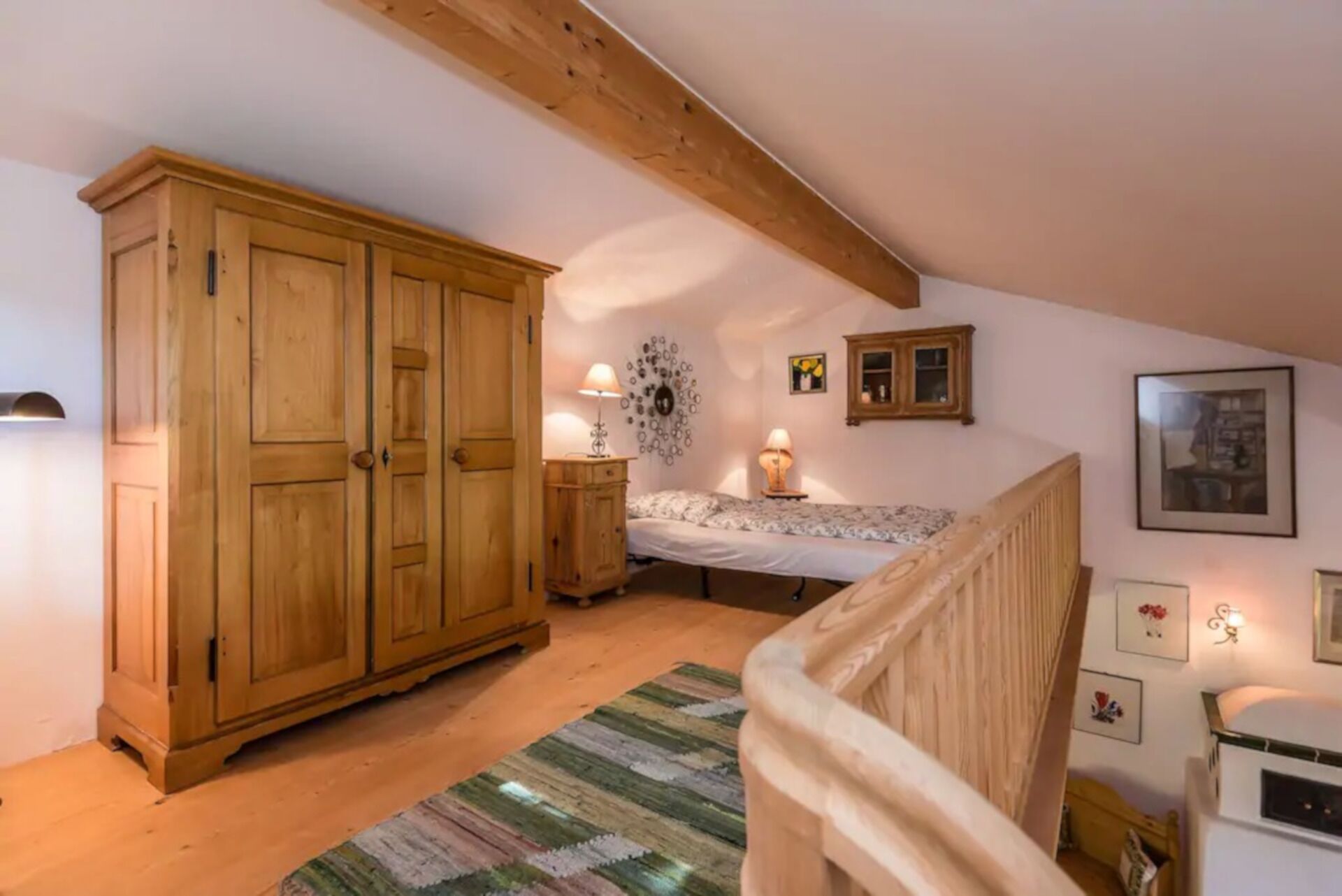 A bedroom with a large wooden wardrobe, a bed with a patterned bedspread, a small bedside table with a lamp, and a decorative wall piece.