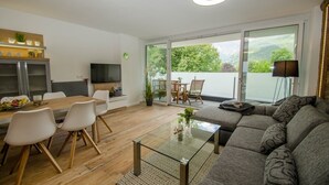 Una sala de estar moderna con un sofá, una mesa de centro de vidrio, una zona de comedor con sillas, un televisor y un balcón con vistas a zonas verdes.