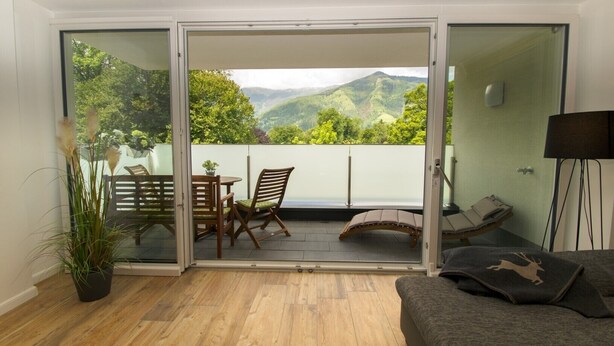 Una habitación con puerta de vidrio corredera que da a un balcón con vistas a colinas verdes. En el interior, hay piso de madera, un sofá y una mesa con sillas.