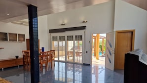 A spacious living area with a large sliding glass door leading to an outdoor patio. There is a wooden table with two remotes on it, and a bench along the wall.