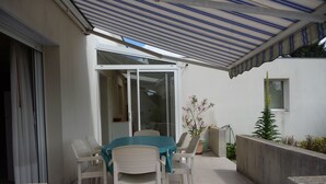 A covered patio with a table and chairs, a sliding glass door, and potted plants. - Gite 37 bis rue du pré de la dame. (Férel)