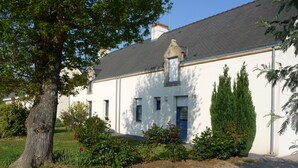 A two-story building with a white facade and dark roof, surrounded by greenery and a gravel driveway. - Gite 37 bis rue du pré de la dame. (Férel)