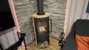 A modern living room with a stone wall, a black wood-burning stove, a wooden cabinet, and a grey sofa with an orange cushion. - Cozy cottage with much comfort (Rheinboellen)
