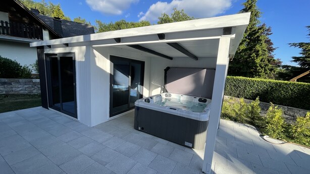 A modern outdoor hot tub area with a pergola overhead, a sliding glass door, and a stone wall in the background. - Cozy cottage with much comfort (Rheinboellen)