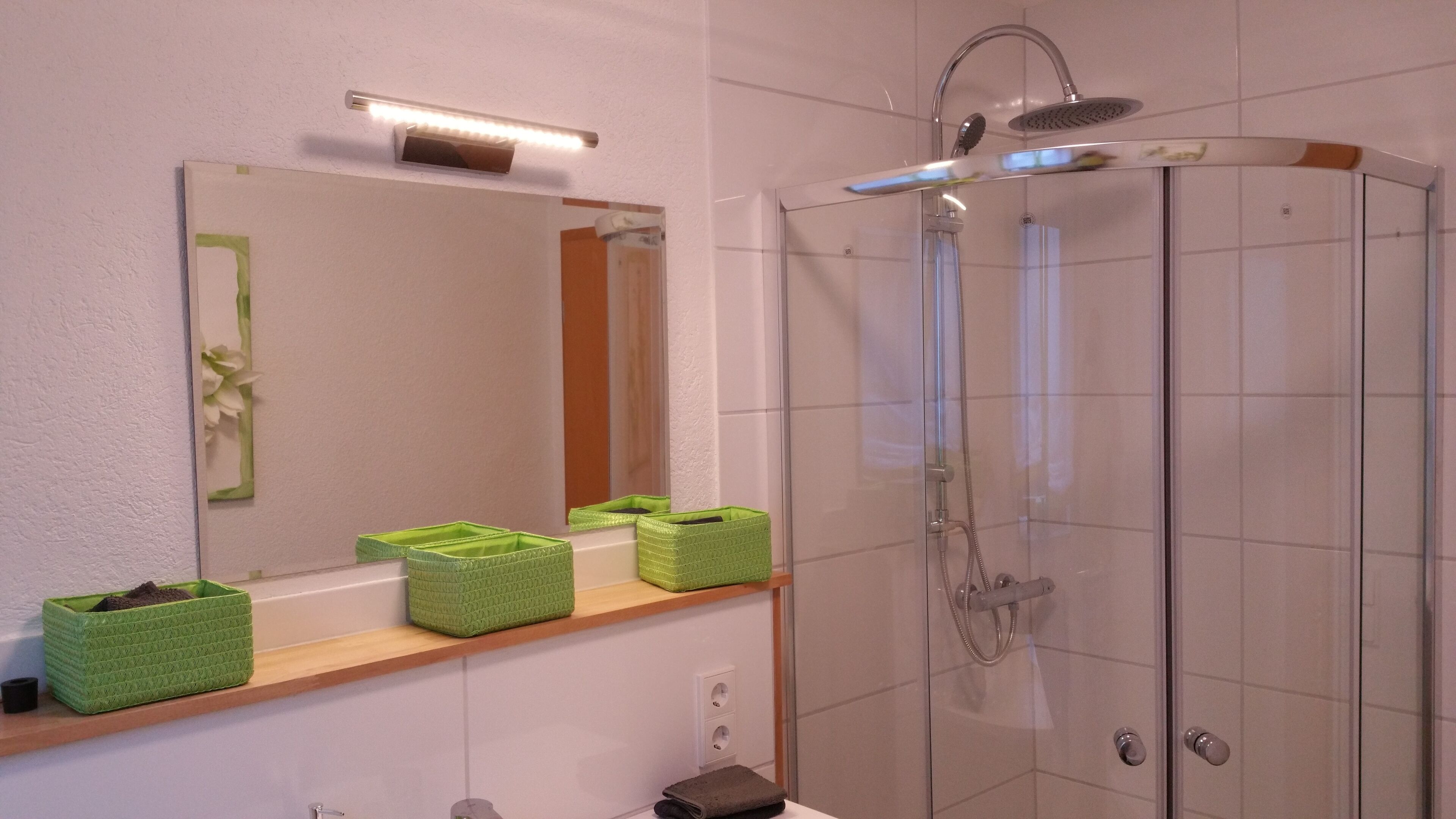 A bathroom with a glass shower enclosure, a mirror with a light above it, and green baskets on a shelf.