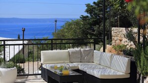 Un balcon avec un canapé blanc, une petite table sur laquelle se trouvent une carafe de jus d’orange et un saladier rempli de fruits, ainsi que des plantes en pot.