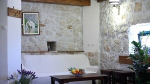 Une pièce aux murs de pierre, avec un canapé blanc, une table en bois sur laquelle se trouve un panier de fruits et une plante en pot.