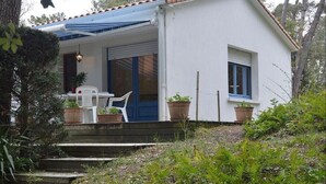 Une maison blanche avec un auvent bleu, une porte blanche et une fenêtre. Des marches mènent à la porte, et des plantes en pot sont disposées sur ces marches.
