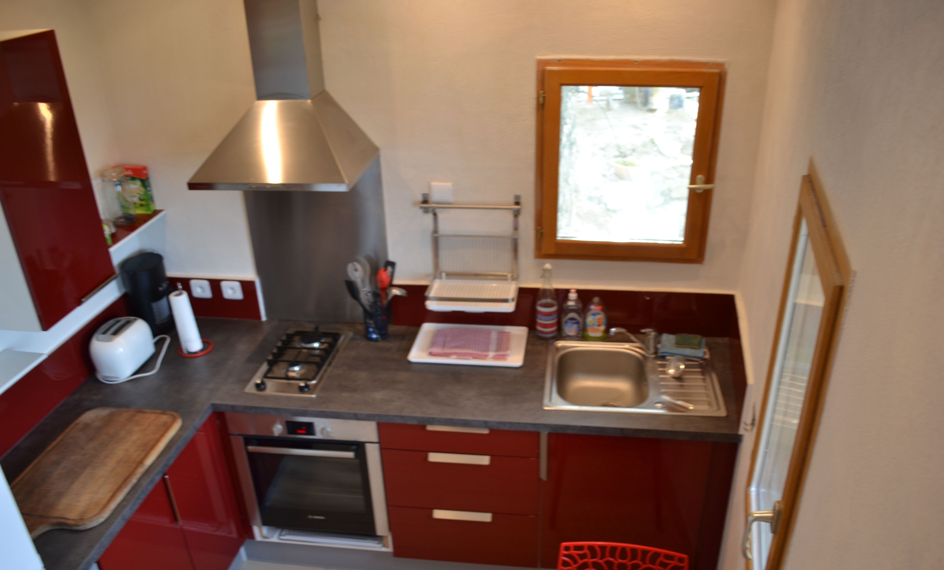Cucina compatta con piano cottura a gas, forno e lavello in acciaio inossidabile. Sopra il lavello c'è una finestra e nell'angolo una sedia rossa.
