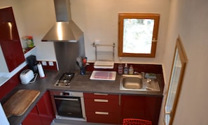 Une cuisine compacte équipée d’une cuisinière à gaz, d’un four et d’un évier en acier inoxydable. Une fenêtre se trouve au-dessus de l’évier et une chaise rouge est placée dans un coin.