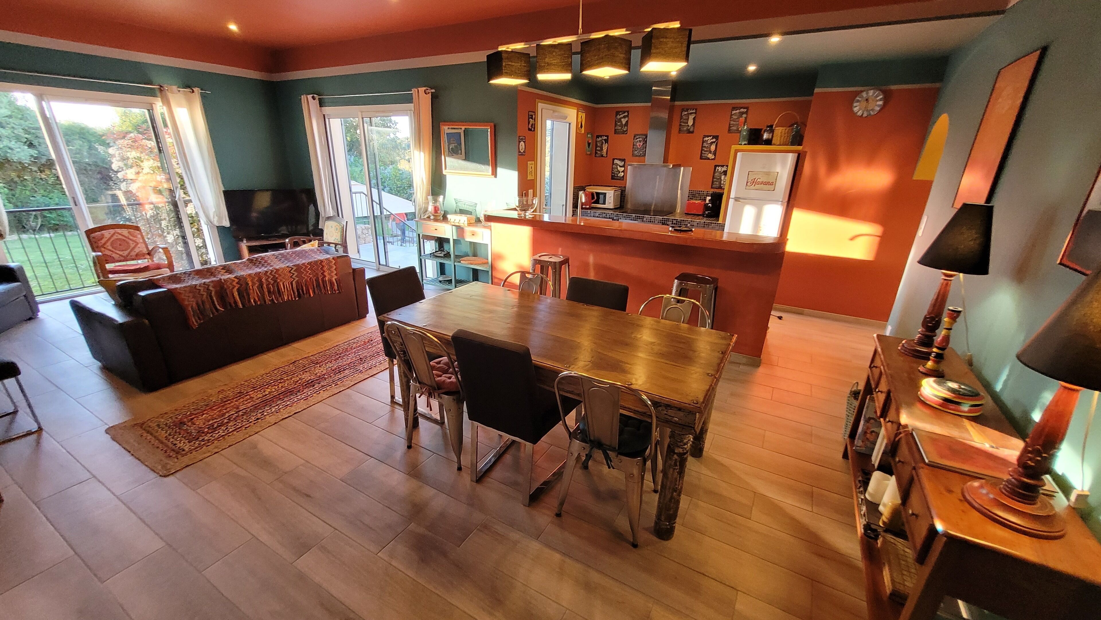 Een eetgedeelte met een houten tafel en stoelen, een bank, en een keuken met een koelkast en magnetron.