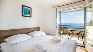 A hotel room with a large bed, two pillows, a wooden headboard, a framed picture on the wall, and a balcony with a table and chairs overlooking the sea. - Villa Marino in Makarska, private pool (Makarska)