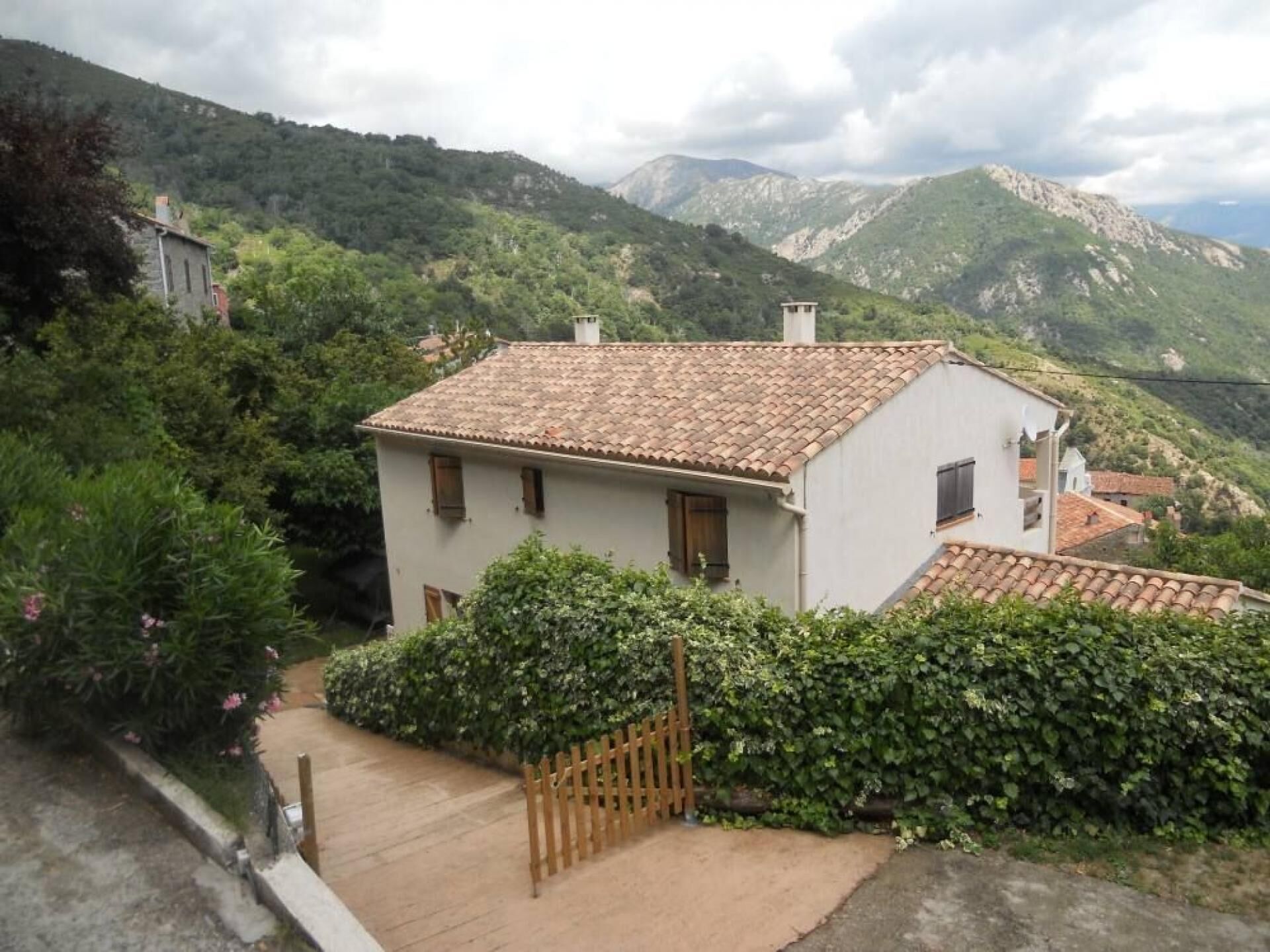 A white building with a tiled roof, surrounded by greenery and mountains in the background.