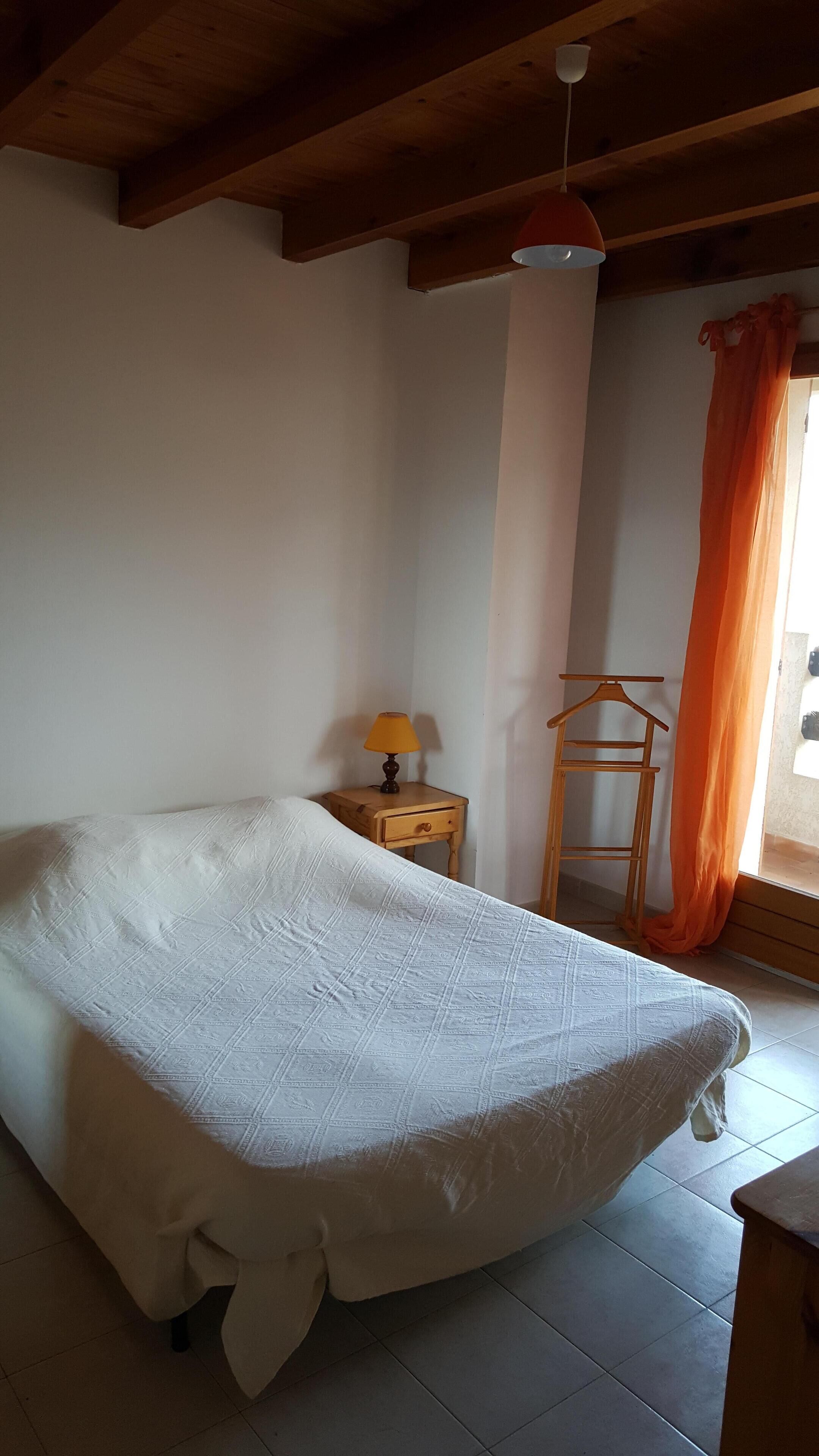 Une chambre à coucher avec un lit, une table de chevet avec une lampe, une chaise en bois et une fenêtre avec des rideaux orange.