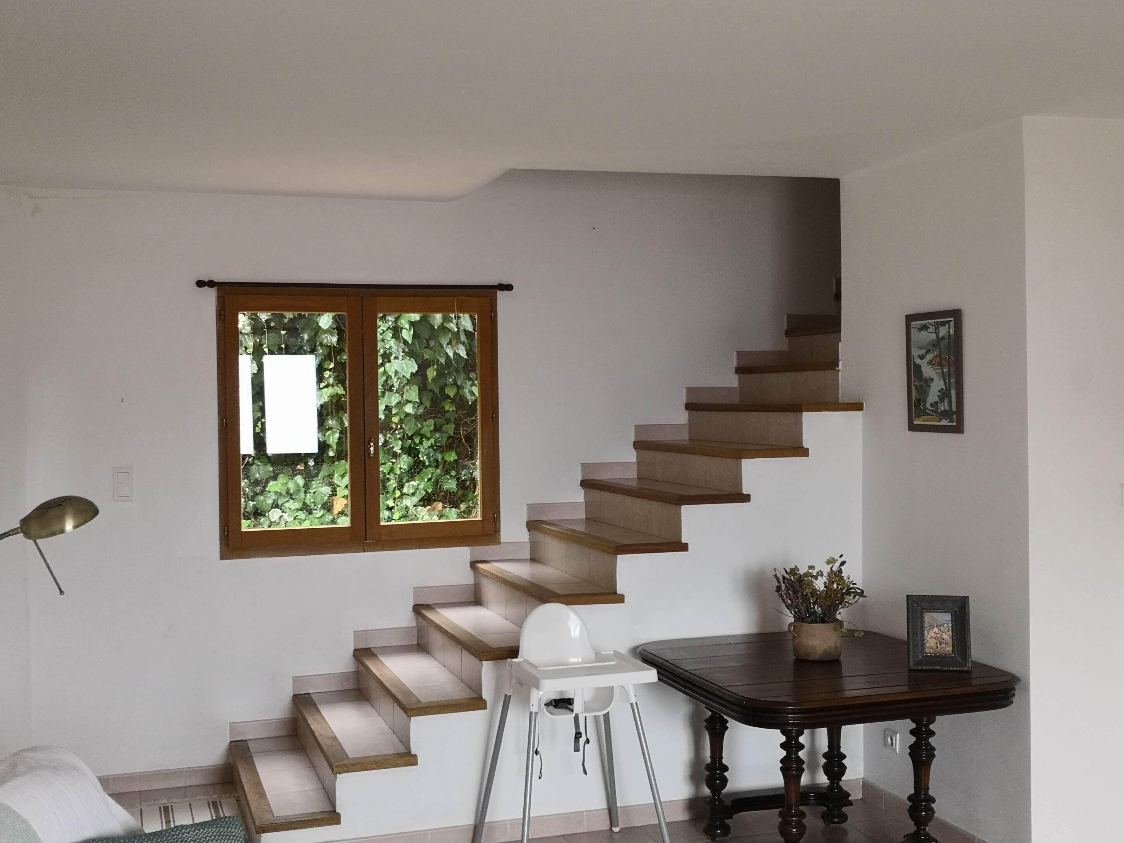 Un escalier avec des marches en bois et une fenêtre donnant sur un paysage verdoyant. On y trouve une table avec un vase et un cadre photo.