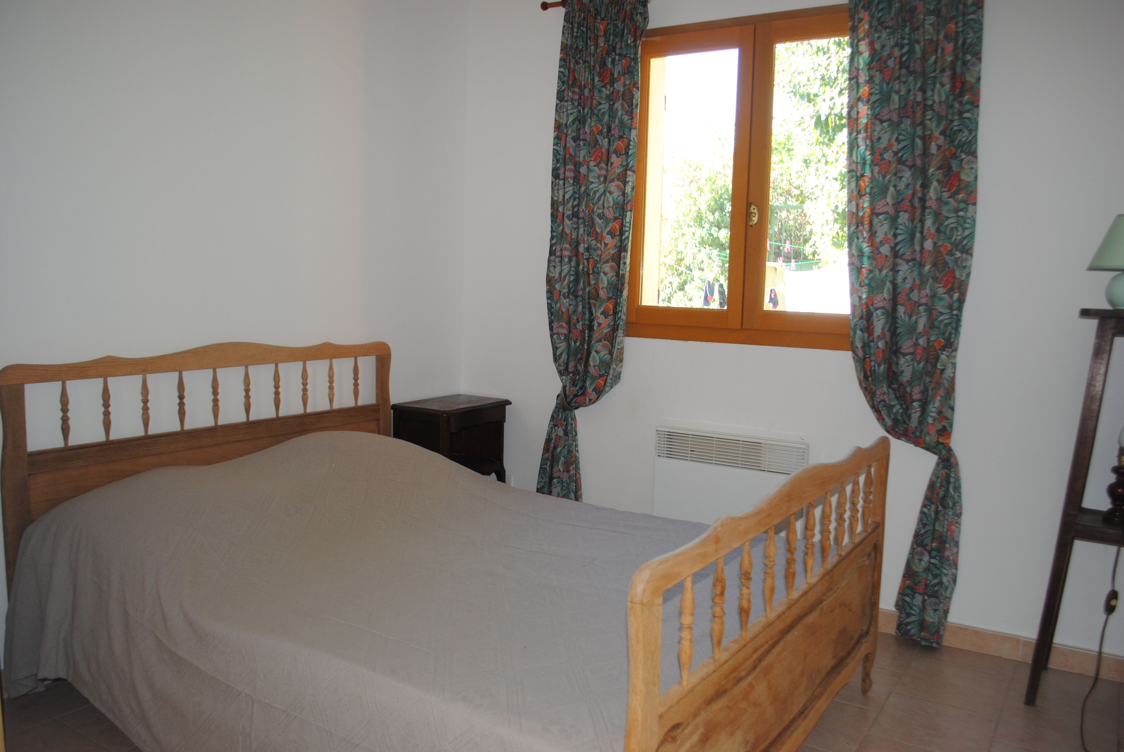 Ein Schlafzimmer mit einem hölzernen Bett, einem Nachttisch, einem Fenster mit Vorhängen und einer Heizung.