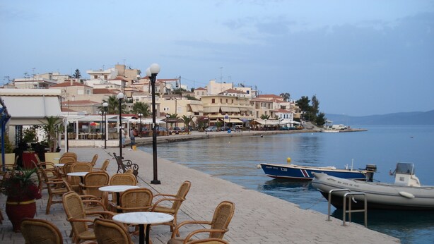 A waterfront café with wicker chairs and tables, a row of buildings in the background, and boats docked at the pier. - Villa KoKa - A small paradise near Ermioni (Petrothalassa Ermioni)