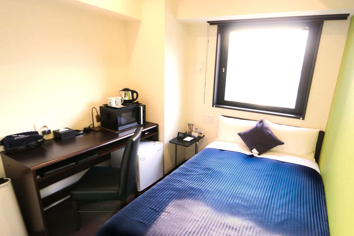 A hotel room with a bed, a desk, a chair, a microwave, a small refrigerator, and a window.
