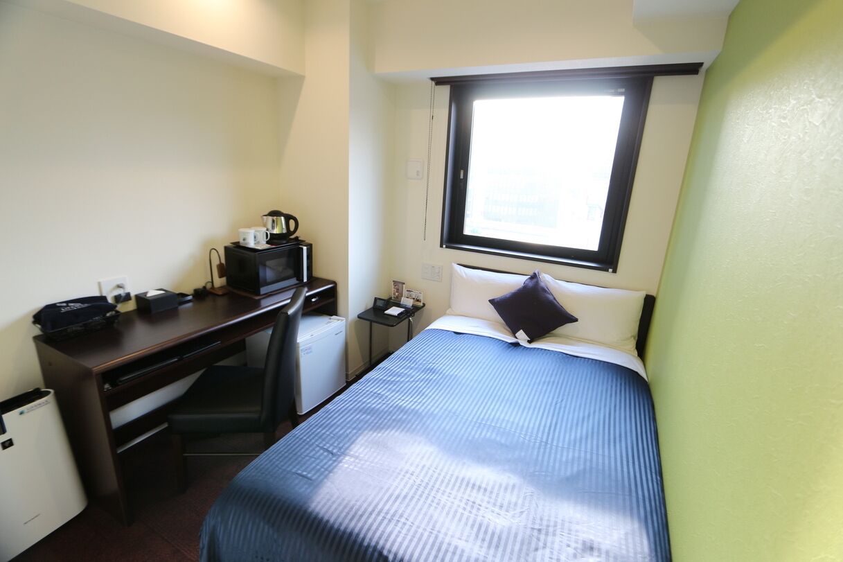 A compact hotel room with a single bed, a desk with a computer, a small refrigerator, and a window providing natural light.