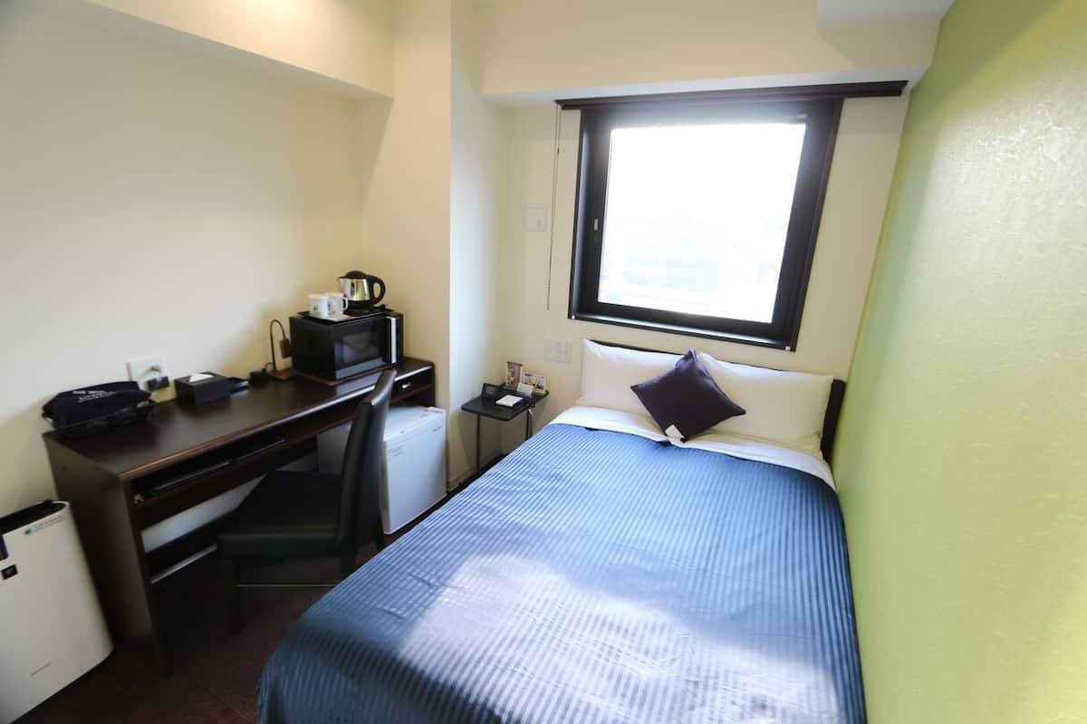 A compact hotel room with a single bed, a desk with a computer, a small refrigerator, and a window providing natural light.
