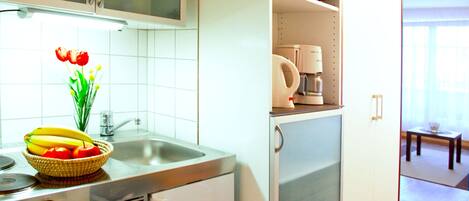A compact kitchen with a microwave, toaster, and a small refrigerator. A sink with a faucet and a bowl of fruit on the counter are also visible.